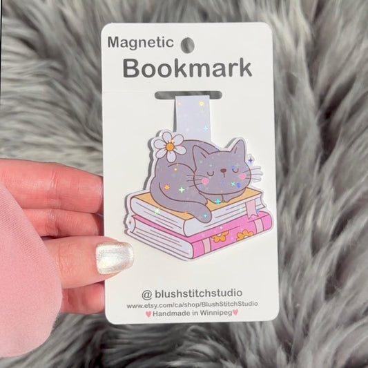 Magnetic bookmark with a cat design on a white card held by a hand against a fluffy gray background.