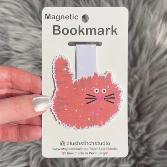 Magnetic bookmark with an orange cat design held by a hand on a fluffy gray surface