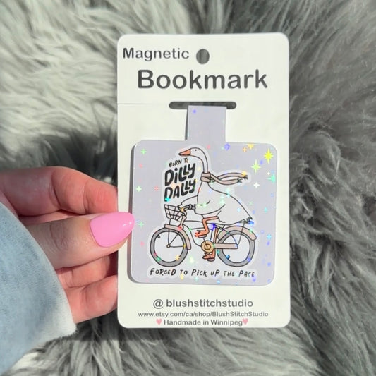 Magnetic bookmark with a cartoon goose on a bike and text, held against a fluffy gray background.