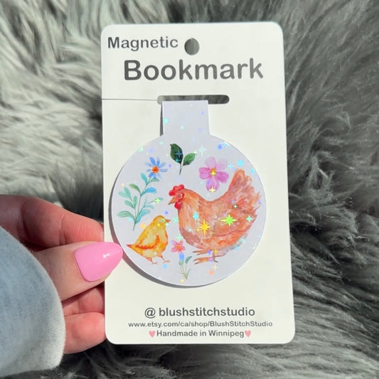 Magnetic bookmark with chicken design on a card held by a hand against a fluffy gray background