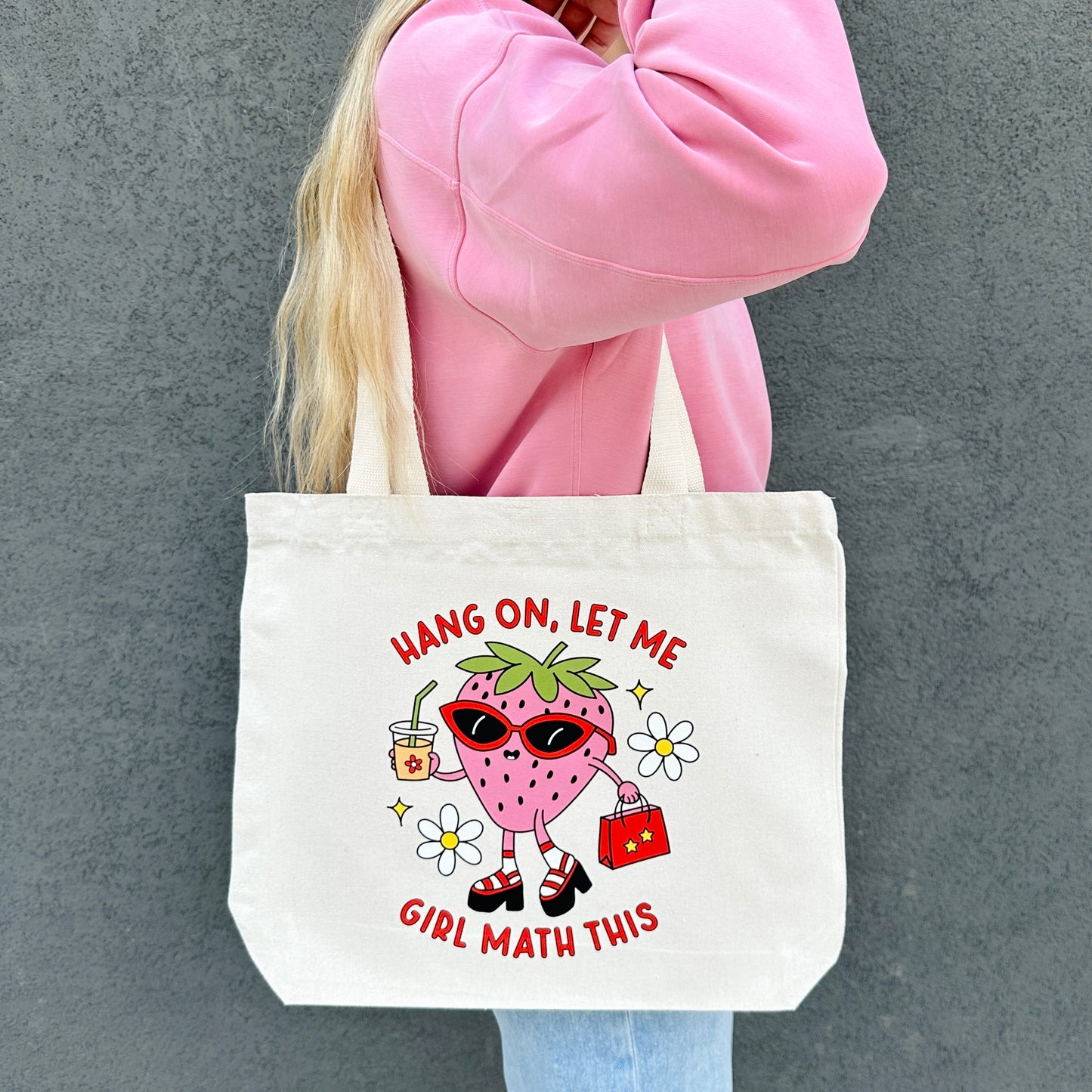 Tote bag with a girl math cartoon strawberry character and text, held by a person in a pink sweatshirt.