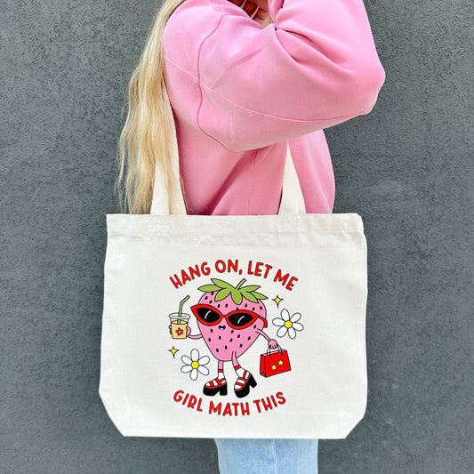 Tote bag with a girl math cartoon strawberry character and text, held by a person in a pink sweatshirt.