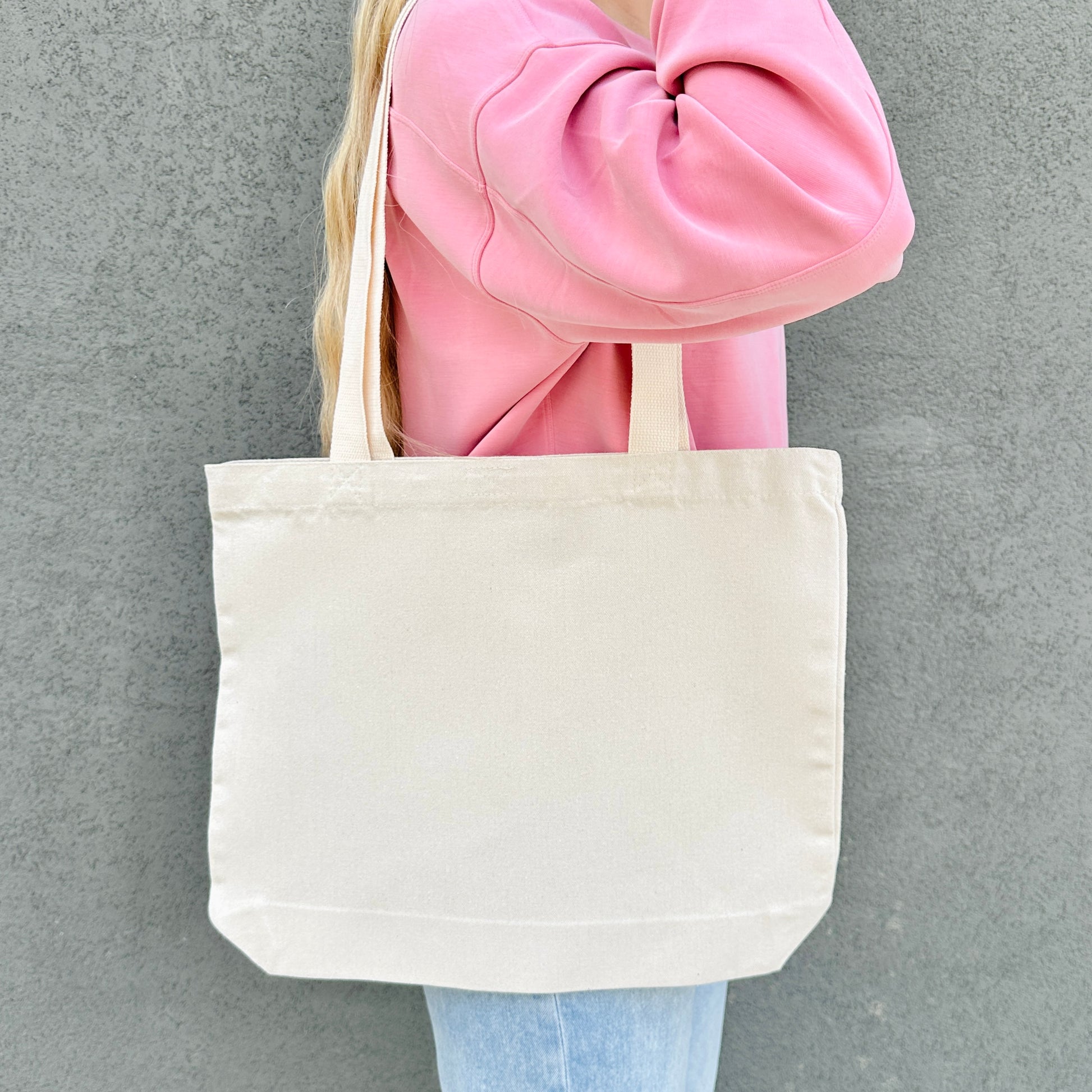 Person holding a beige tote bag against a gray background. the backside of the tote bag does not have a design. 