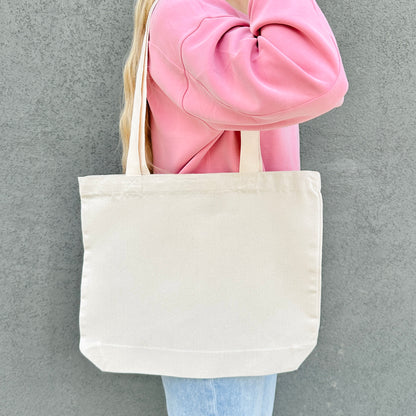 Person holding a beige tote bag against a gray background. the backside of the tote bag does not have a design. 
