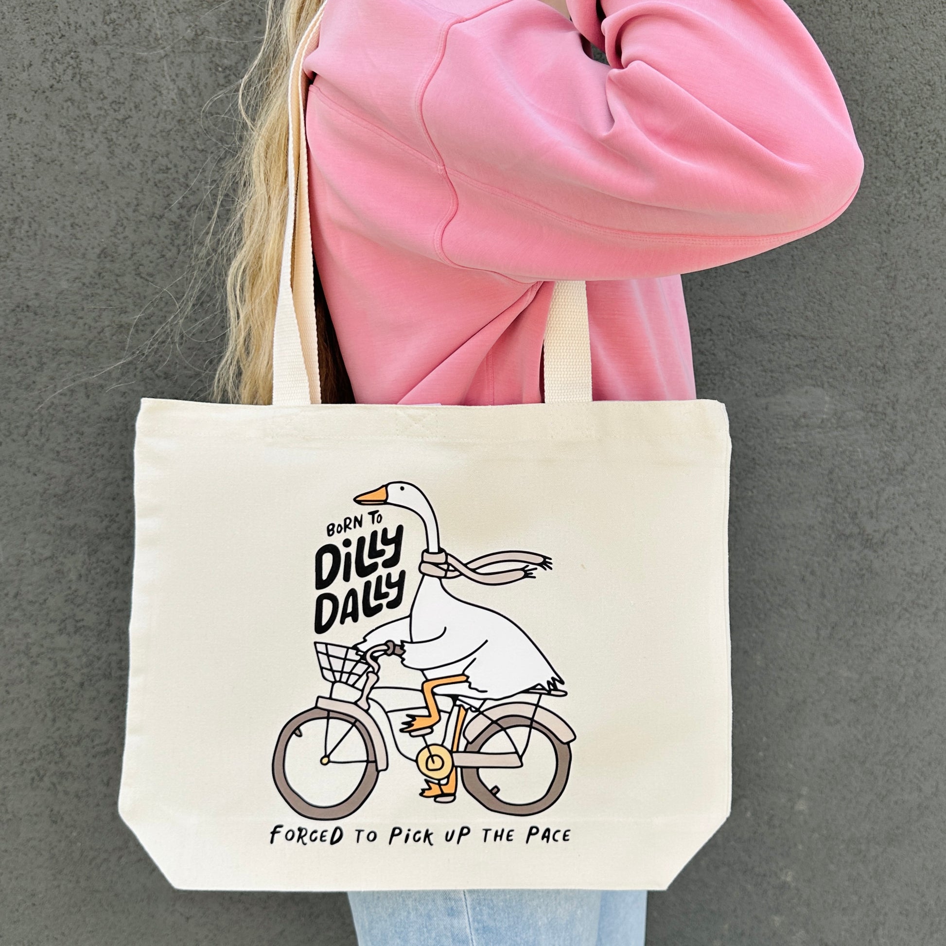 Tote bag with a born to dilly dally goose design , held by a person in a pink jacket.
