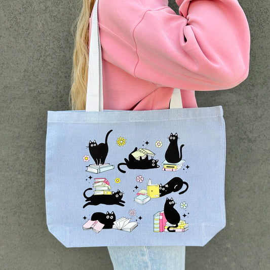 blue tote bag with black cat and books illustrations held by a person wearing a blue sweater.