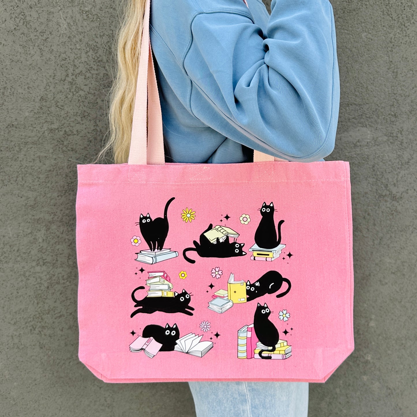 Pink tote bag with black cat and books illustrations held by a person wearing a blue sweater.