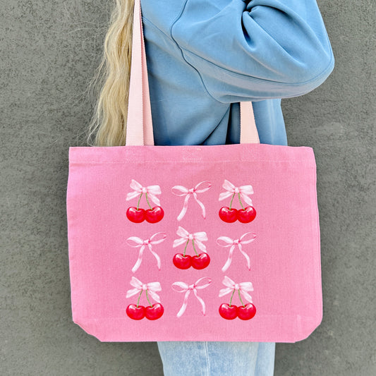 Pink tote bag with cherry and bow design held by a person against a gray background