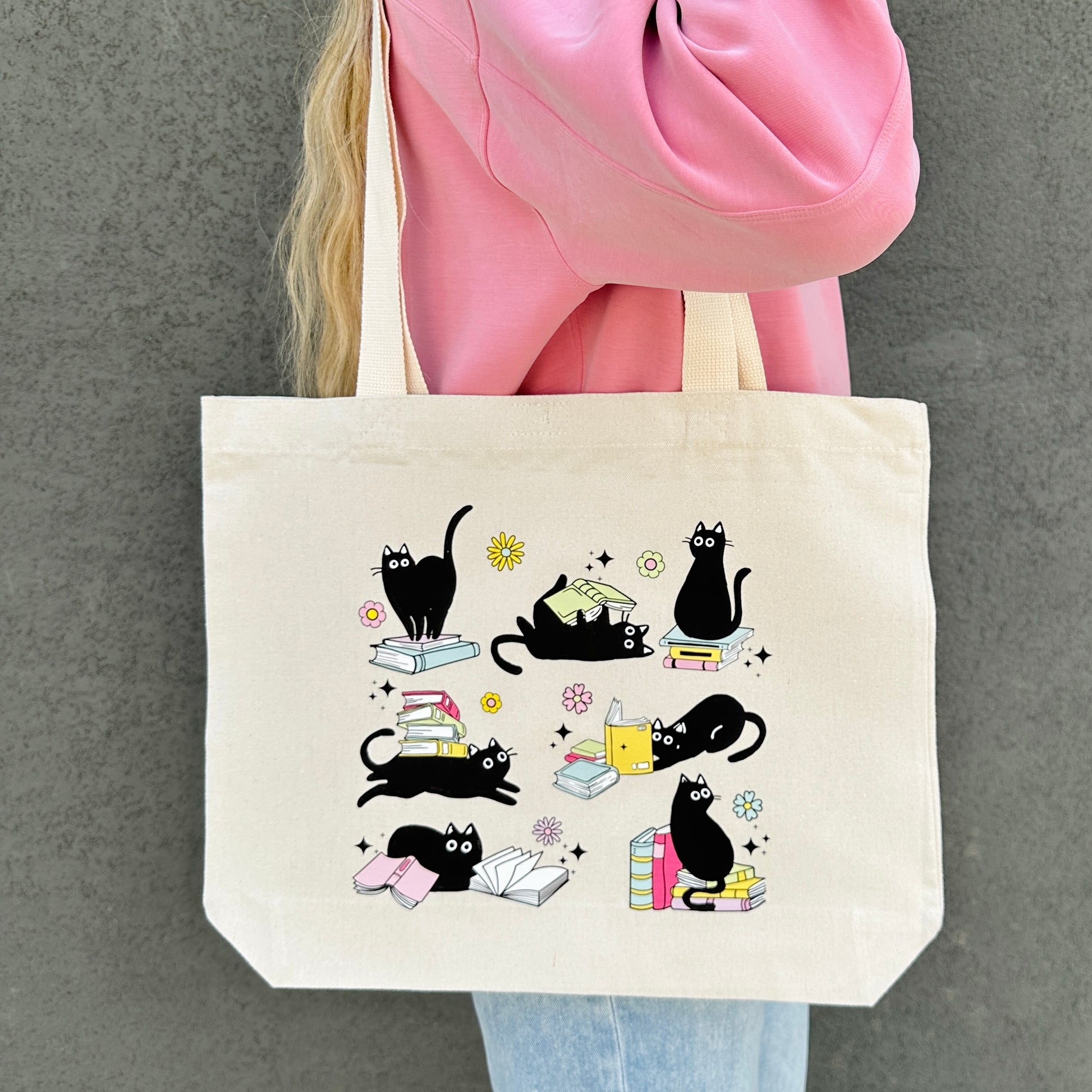 Tote bag with black cat and book illustrations held by a person wearing a pink jacket.