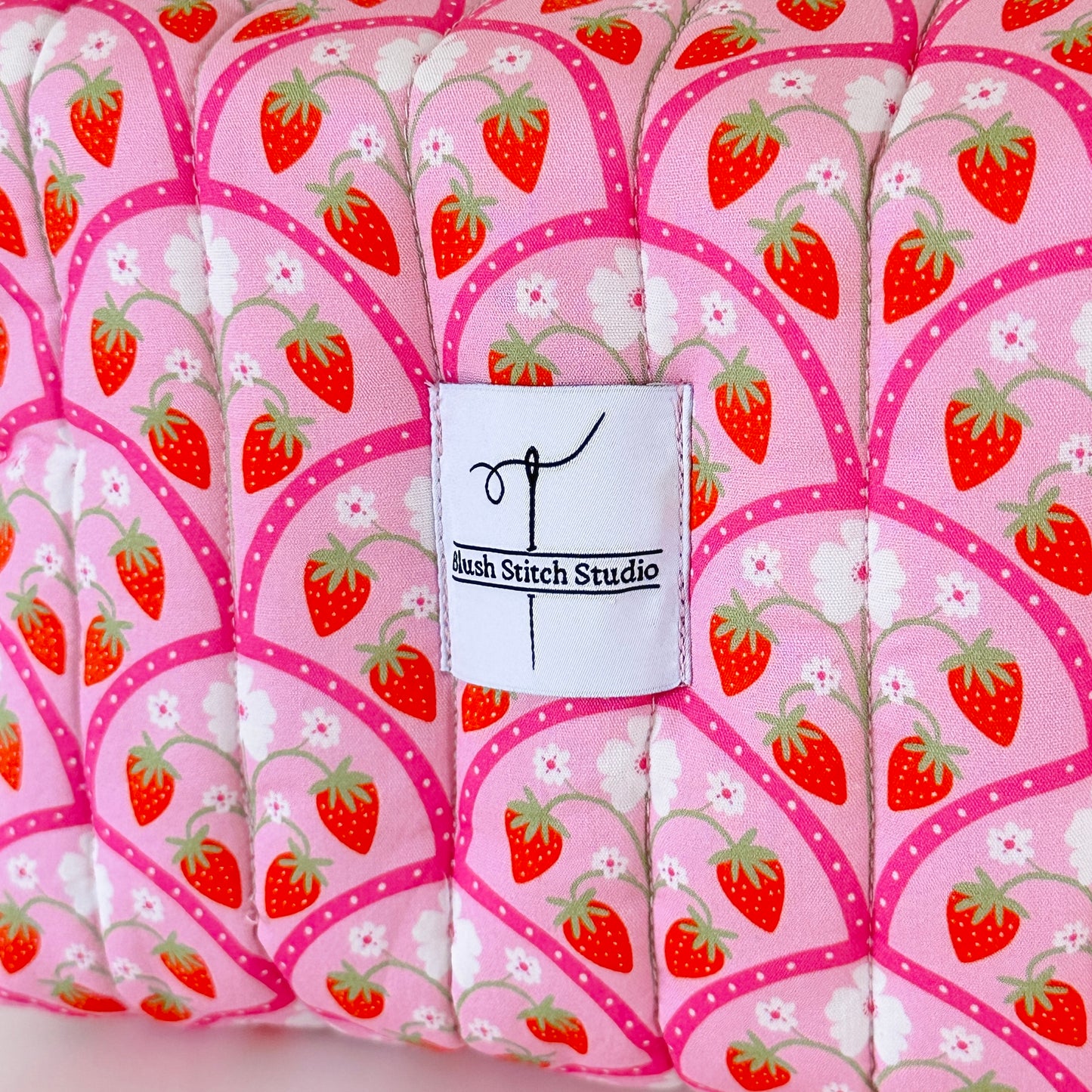 Fabric with strawberry pattern and a label for Bush Stitch Studio on a pink background