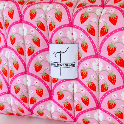 Fabric with strawberry pattern and a label for Bush Stitch Studio on a pink background