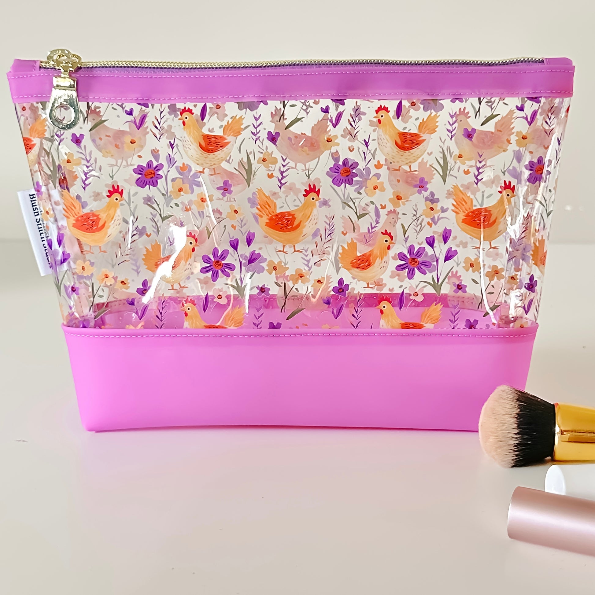 Clear makeup bag with chicken and floral pattern on a pink base, with makeup brush and container on a light background