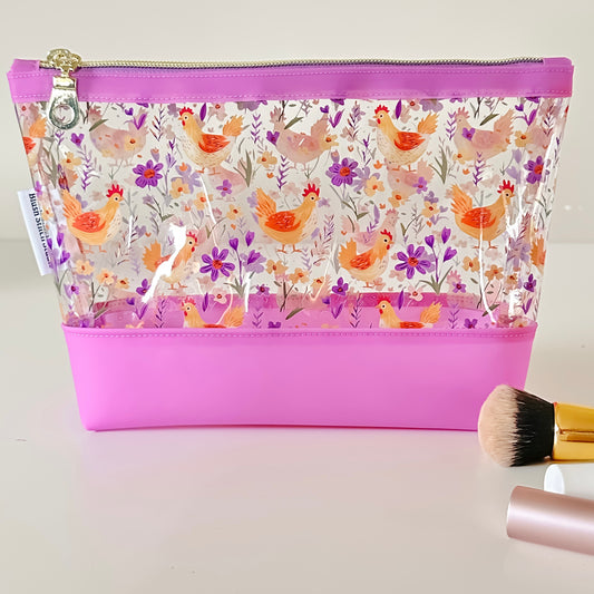 Clear makeup bag with chicken and floral pattern on a pink base, with makeup brush and container on a light background