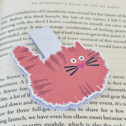 Magnetic bookmark with an orange cat design held by a hand on a fluffy gray surface