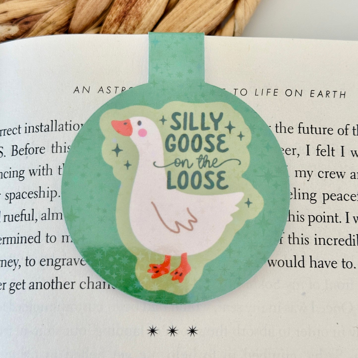 Round magnetic bookmark with a goose illustration and text 'Silly Goose on the Loose' on a book page.