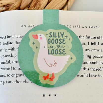 Round magnetic bookmark with a goose illustration and text 'Silly Goose on the Loose' on a book page.