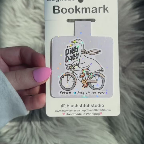 Magnetic bookmark with a cartoon goose on a bike and text, held against a fluffy gray background.