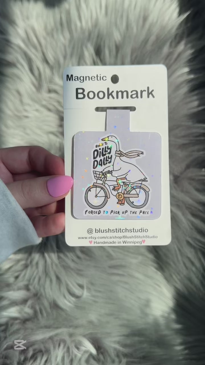 Magnetic bookmark with a cartoon goose on a bike and text, held against a fluffy gray background.