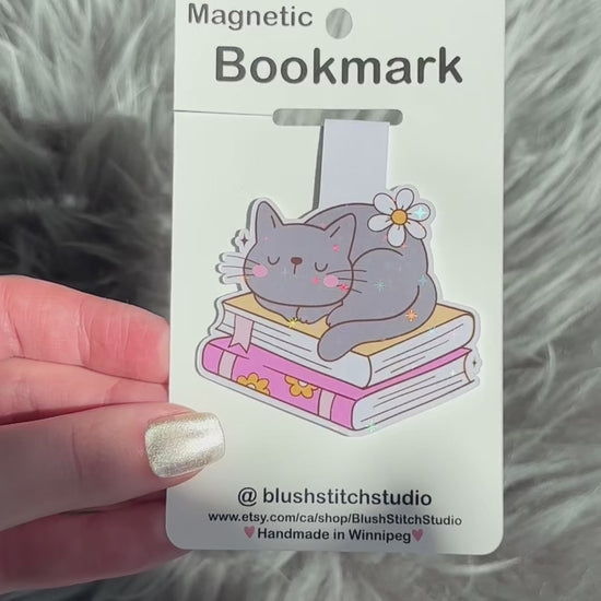 Magnetic bookmark with a cat design on a white card held by a hand against a fluffy gray background.