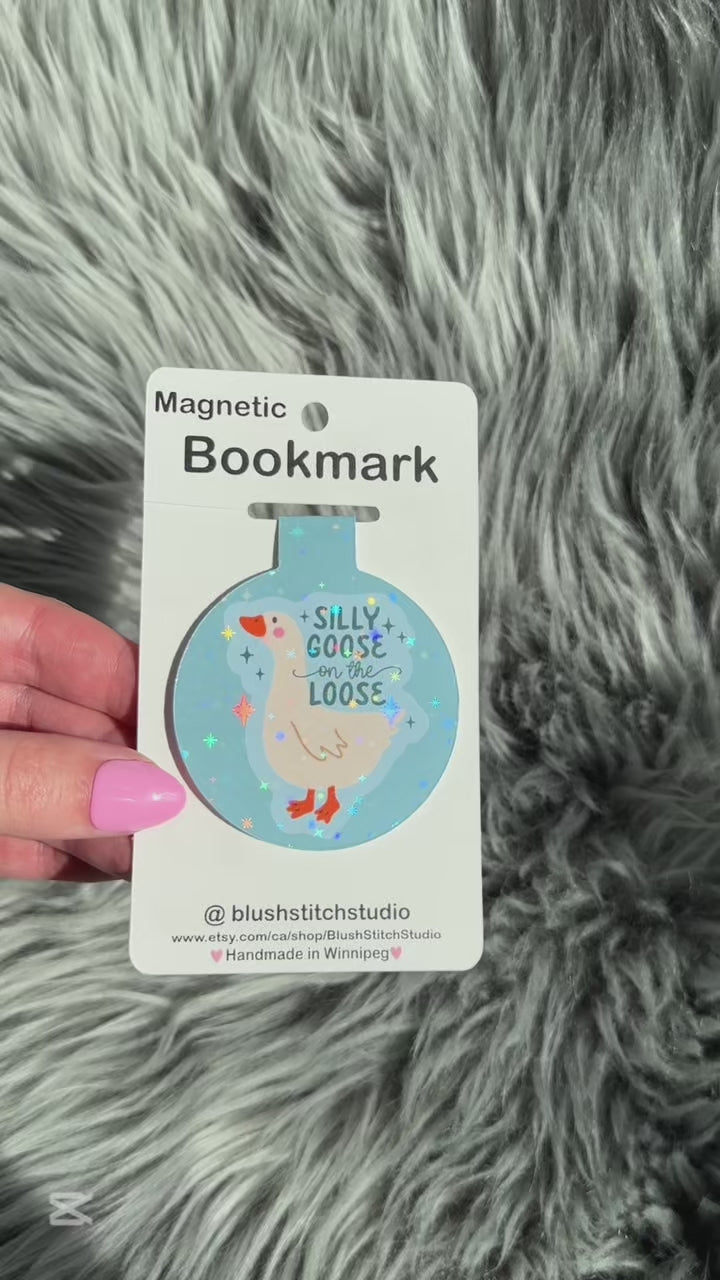 Magnetic bookmark with a silly goose design on a card held by a hand against a fluffy gray background