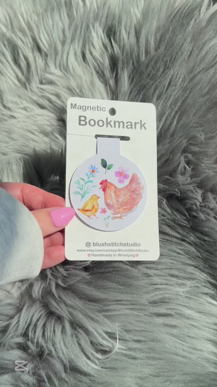 Magnetic bookmark with chicken design on a card held by a hand against a fluffy gray background