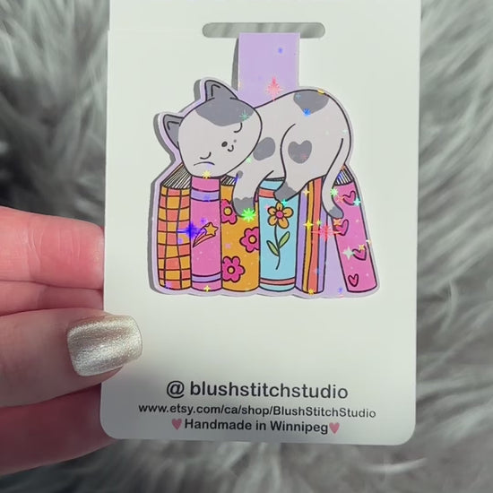Magnetic bookmark with cat design on a Blush Stitch Studio card, held against a fluffy gray background.