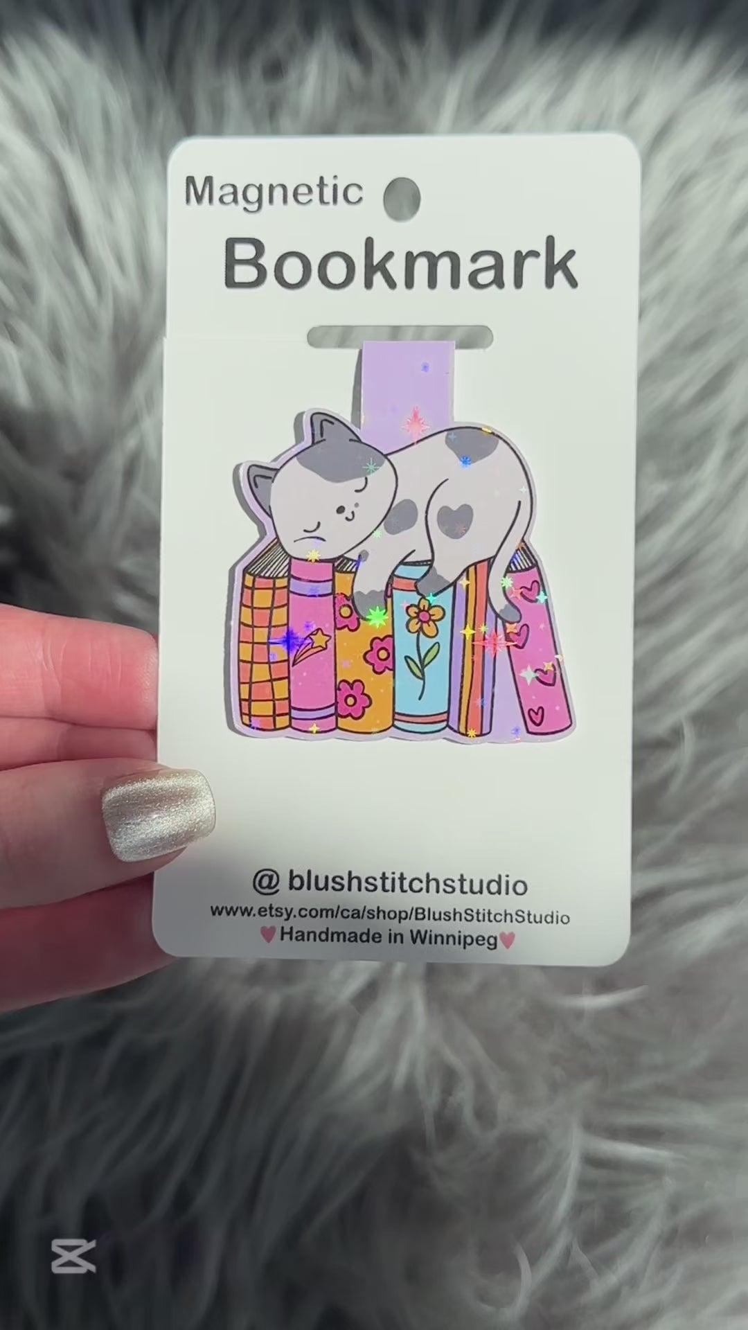 Magnetic bookmark with cat design on a Blush Stitch Studio card, held against a fluffy gray background.