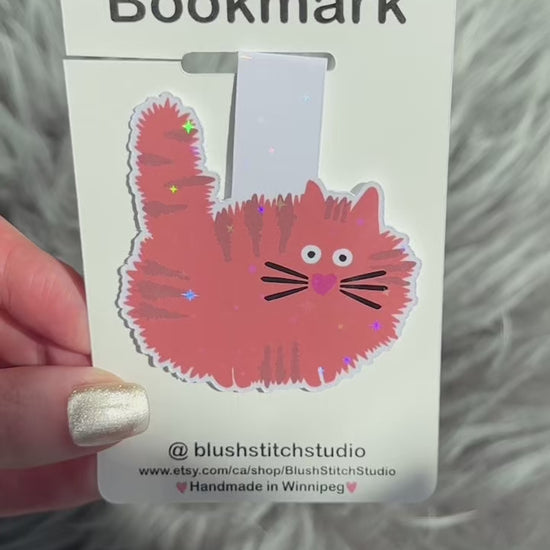 Magnetic bookmark with an orange cat design held by a hand on a fluffy gray surface