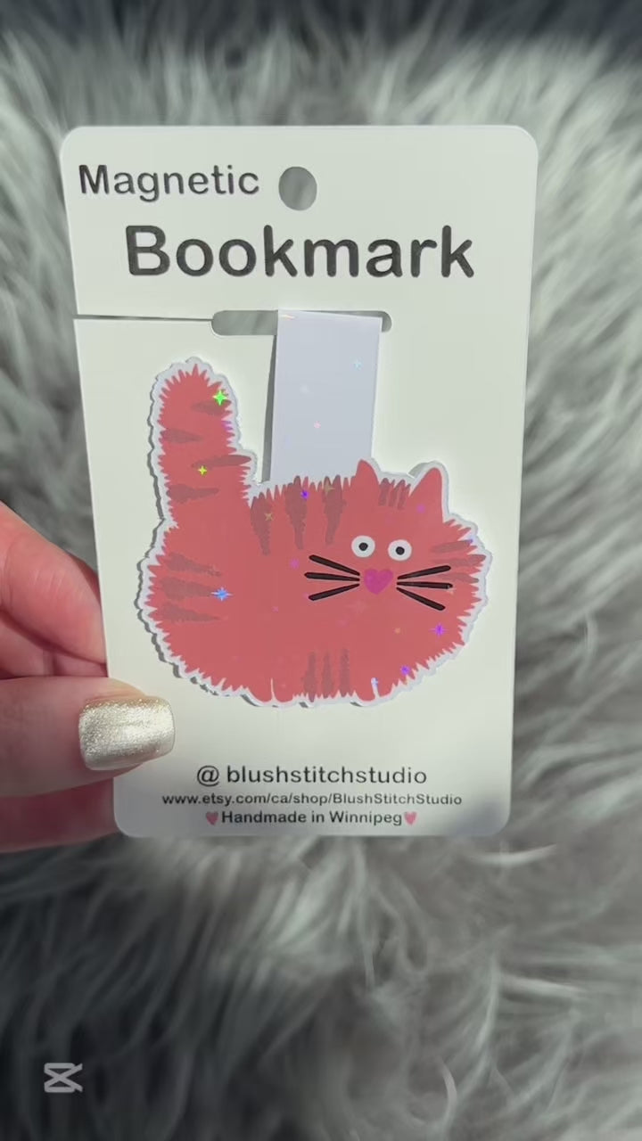 Magnetic bookmark with an orange cat design held by a hand on a fluffy gray surface