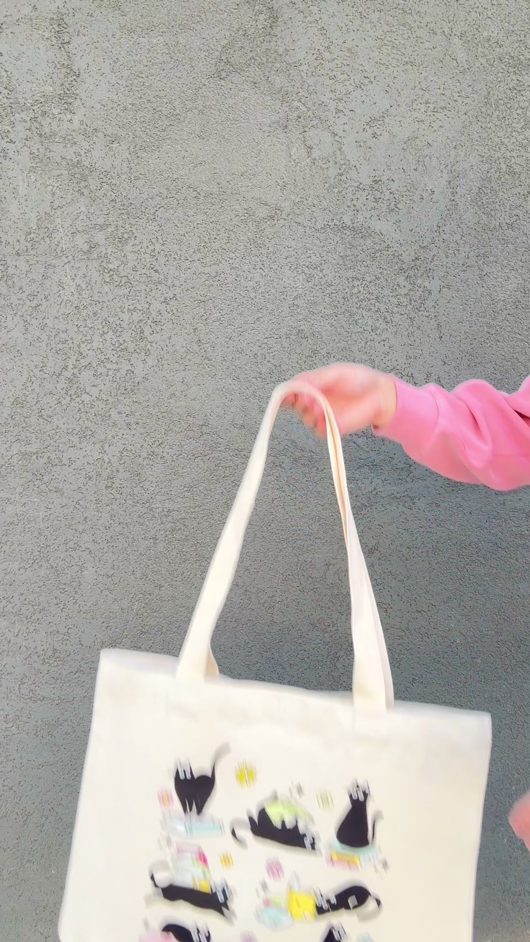Tote bag with black cat and book illustrations held by a person wearing a pink jacket.