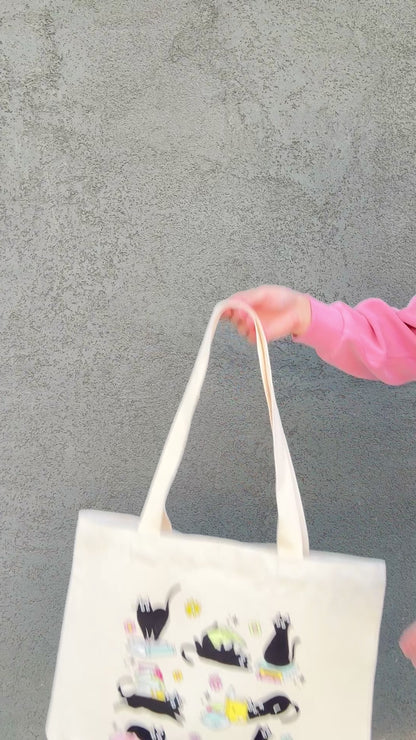 Tote bag with black cat and book illustrations held by a person wearing a pink jacket.
